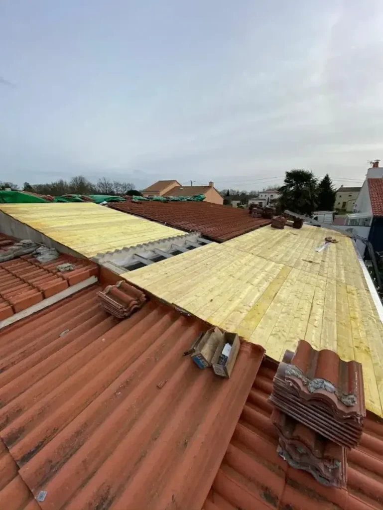 Rénovation de Toiture : Pose de Planches de Bois sur Tuiles Rénovation de toiture avec pose de planches de bois sur des tuiles rouges ondulées sous un ciel nuageux.